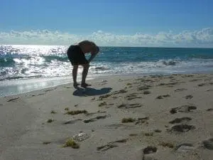 Person bending over on a sandy beach with the ocean in the background.