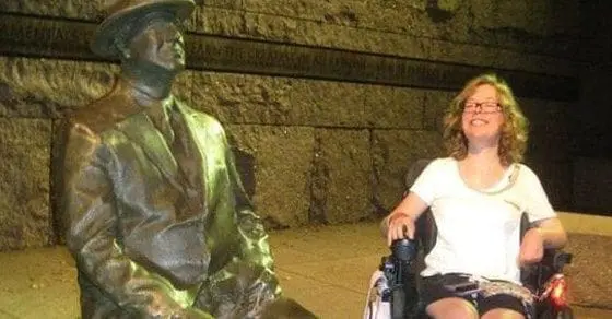 A woman in a wheelchair smiles next to a statue of a man in a hat.