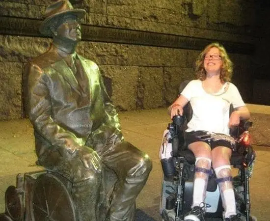 Lisa Lupi smiling while sitting in a wheelchair next to a statue of Franklin D. Roosevelt.