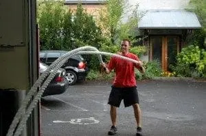 Man in red shirt and black shorts using battling ropes outdoors.