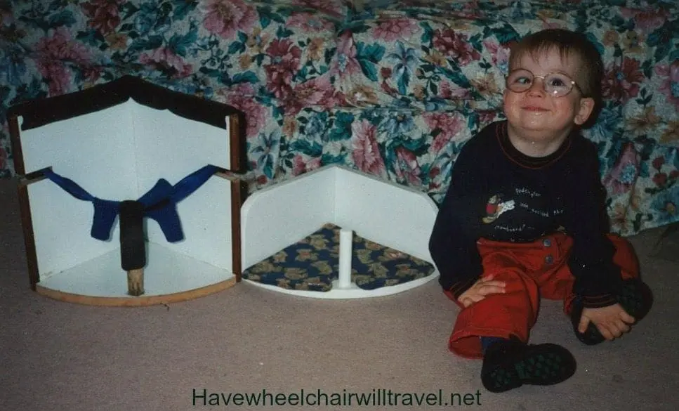 BJ, a young boy wearing glasses, red pants, and a dark blue shirt, sits on the floor next to two pieces of white seating equipment.