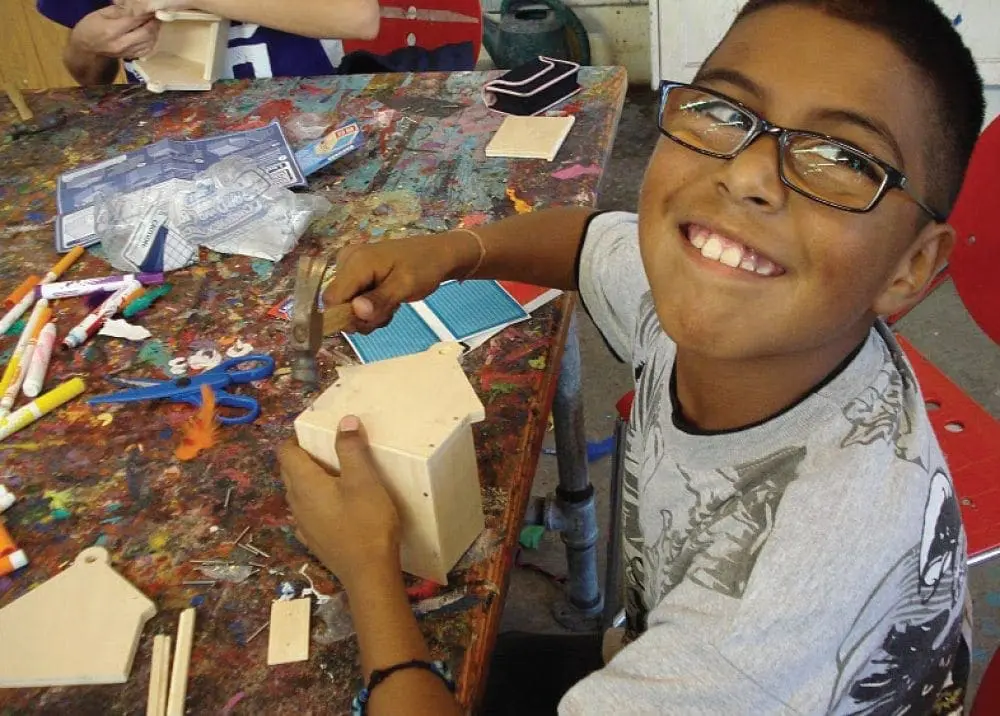 Young boy with glasses smiling while crafting a wooden house.
