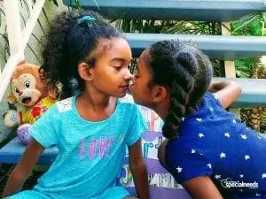 Two young sisters lean in for a kiss, a stuffed monkey to their left.