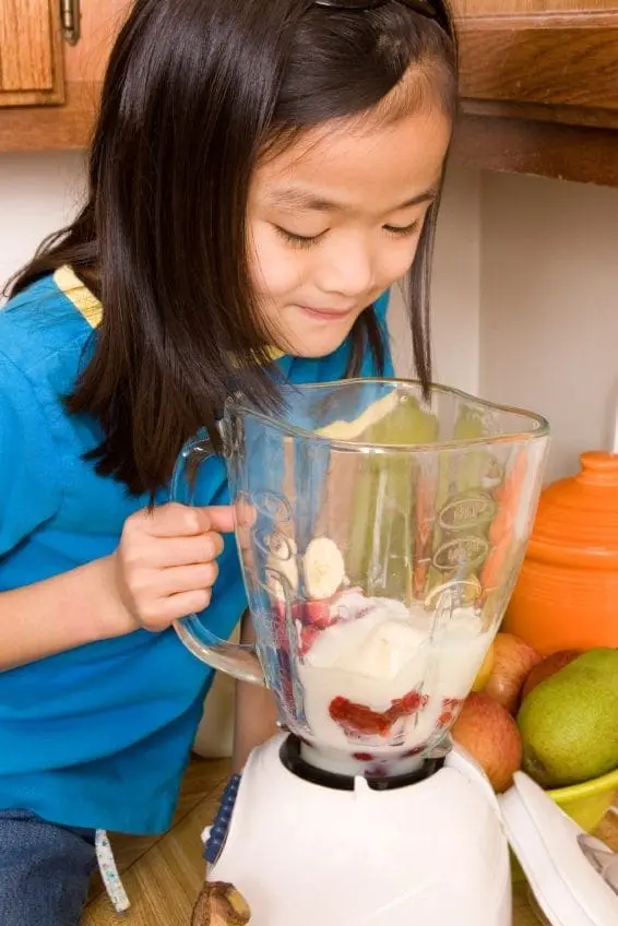 A young girl looks down at a blender filled with fruit and other ingredients.