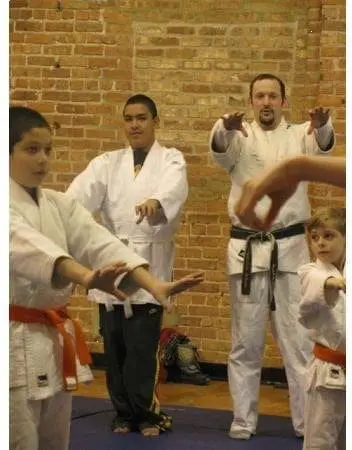 Two men and two children in martial arts uniforms practice stances in front of a brick wall.