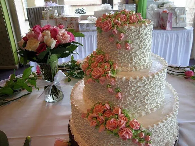 A three-tiered white cake adorned with pink roses and ruffled frosting.
