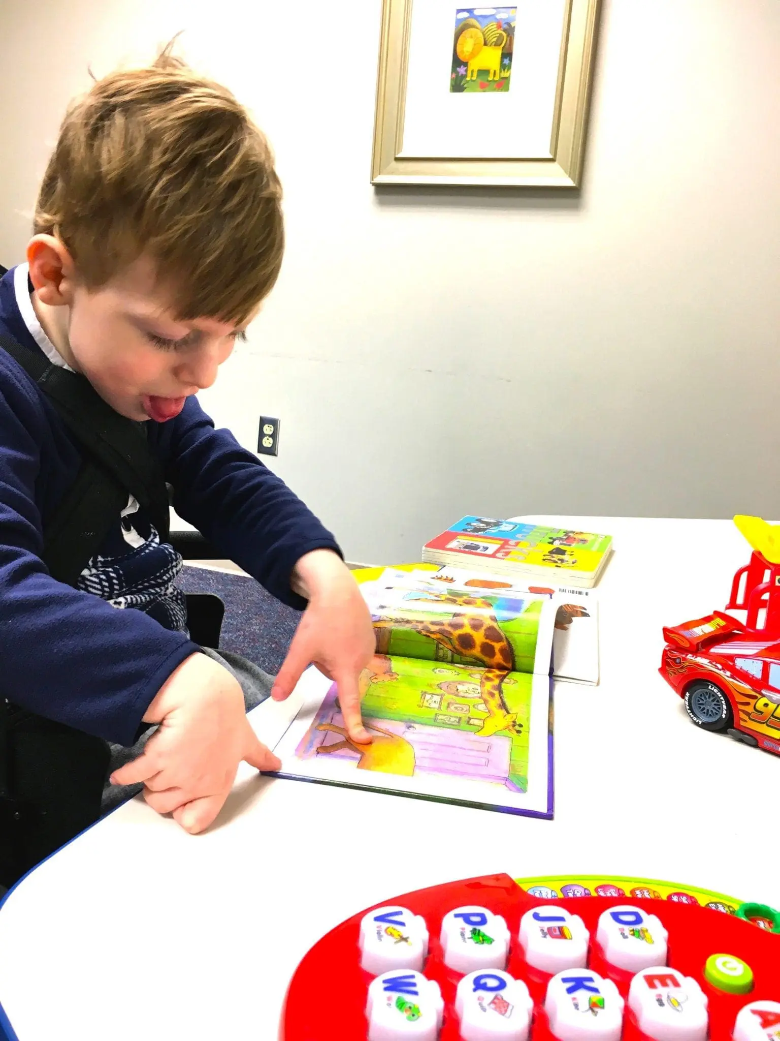 Boy pointing at a children's book with a toy car and alphabet toy on the table.