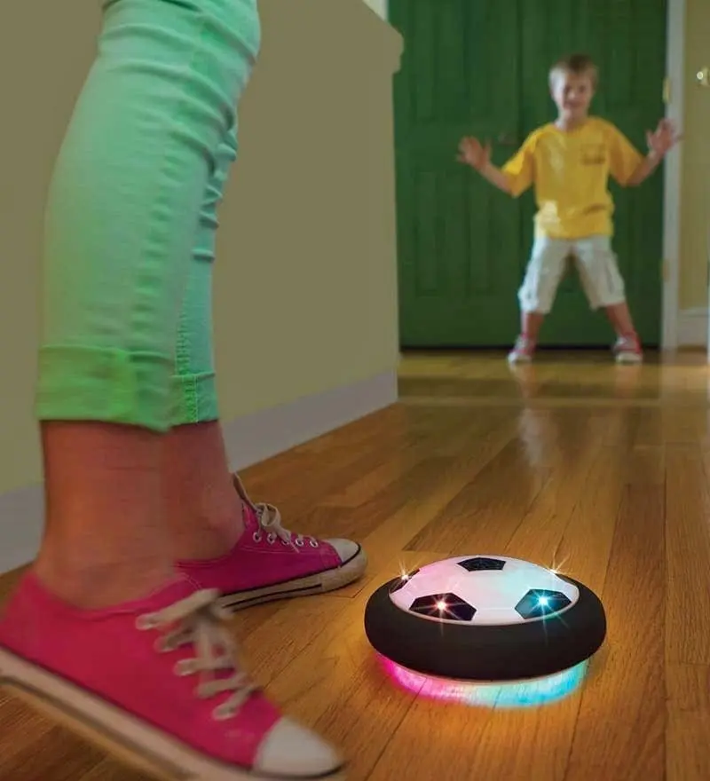 A boy in a yellow shirt plays with an air power soccer disk with a person in pink sneakers and green pants.