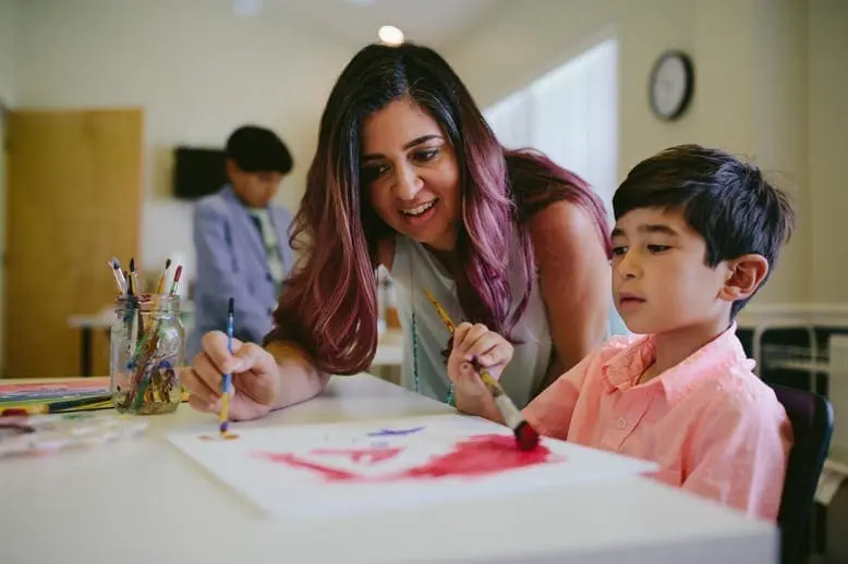 A woman with purple-streaked hair guides a young boy as he paints.