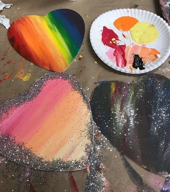 Four heart-shaped art projects, one with rainbow stripes, one with glitter and pink-to-orange paint, one with black glitter, and a plate of paints.