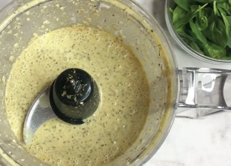 Cashew cream sauce in a food processor with basil in a bowl to the right.