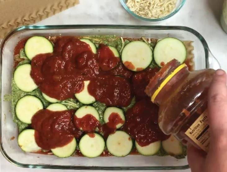 Hand pours tomato sauce onto layers of zucchini and cashew cream in a baking dish.