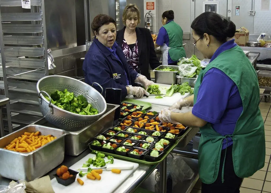 Three people in a kitchen preparing food, including chopped vegetables and a tray of food portions.