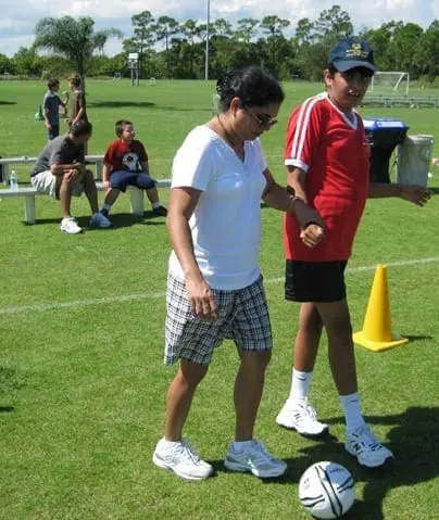 A woman and a boy walk across a soccer field with a soccer ball nearby.