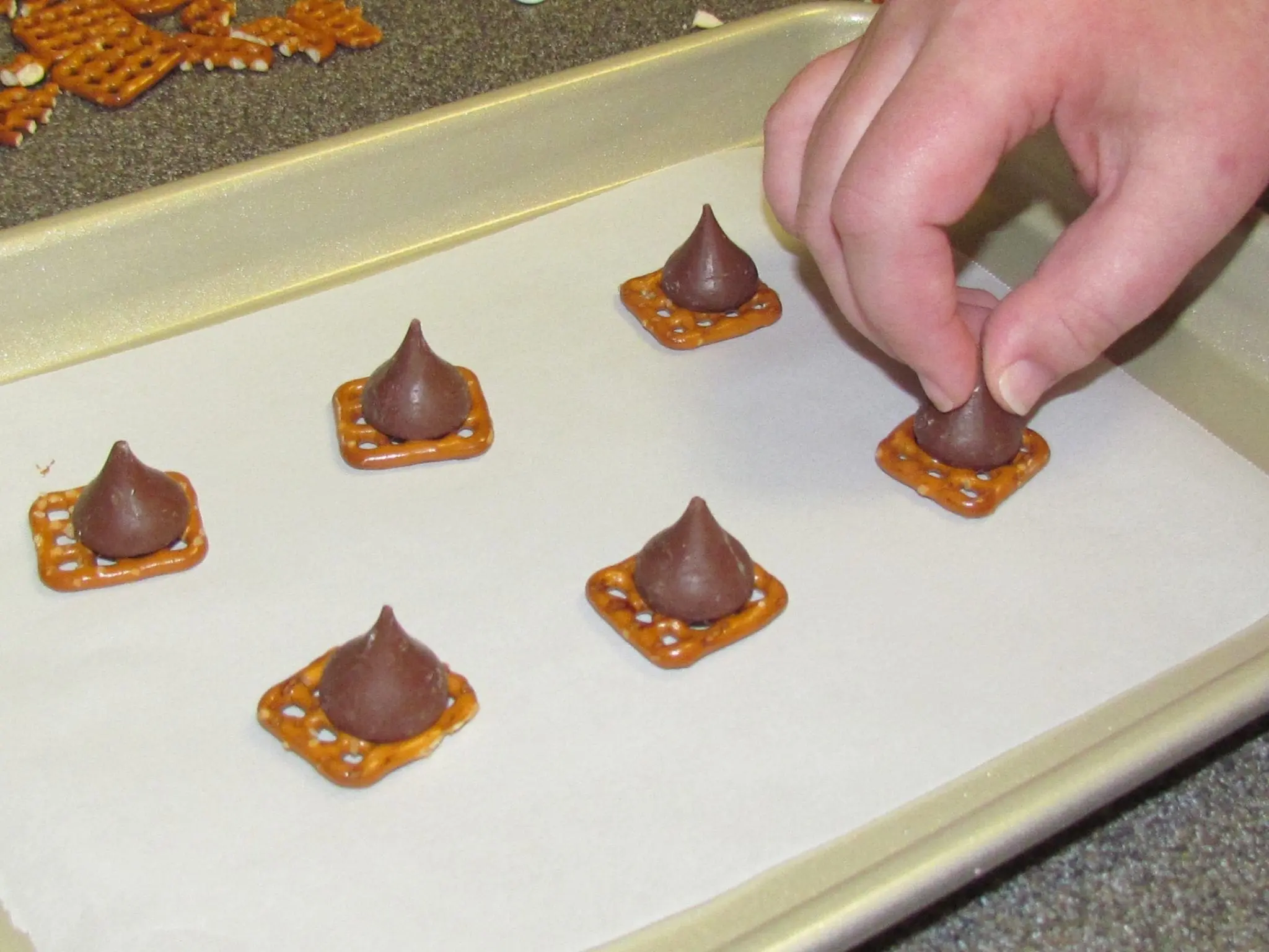 A hand places a chocolate kiss on a pretzel on a baking sheet.