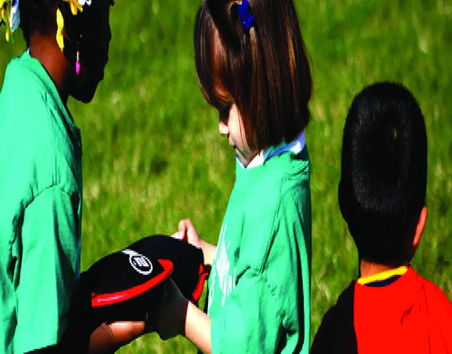 Children in matching green shirts examining a sandbell on a grassy field.