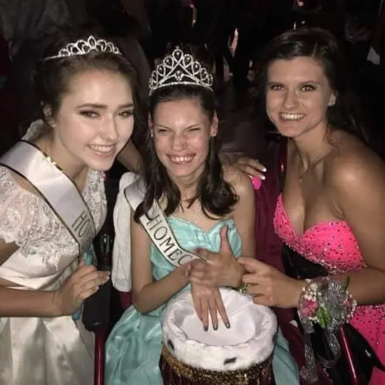 Three young women wearing crowns and sashes, one in a wheelchair, the center woman is crowned Homecoming Queen.