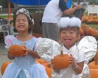 Two young girls in costumes hold pumpkins at a pumpkin patch.