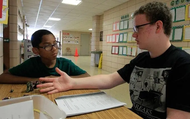 Two young men sitting at a table with papers and objects, one gesturing with his hand.