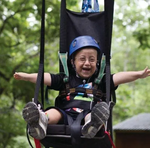 Child in a helmet and harness smiling in a swing outdoors.