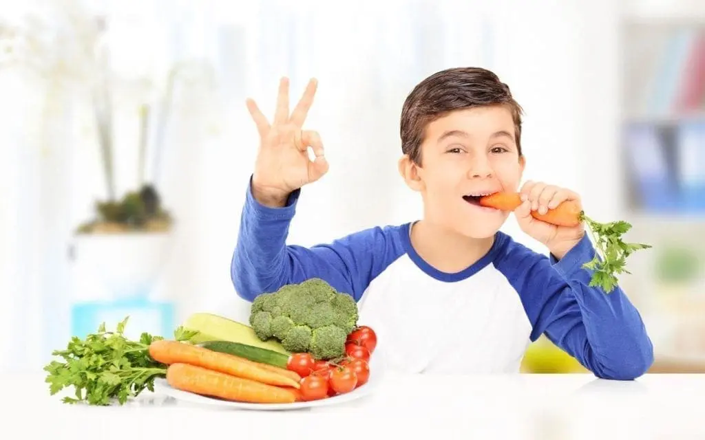 Boy eating a carrot and giving an OK sign with a plate of fresh vegetables in front of him.