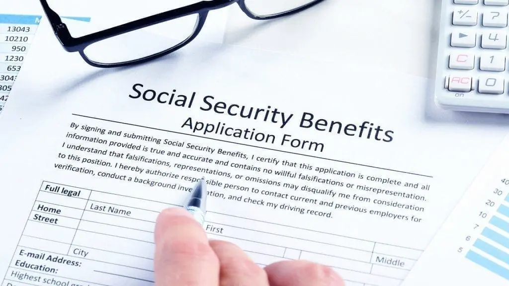 Hand holding a pen over a Social Security Benefits application form with glasses and a calculator nearby.