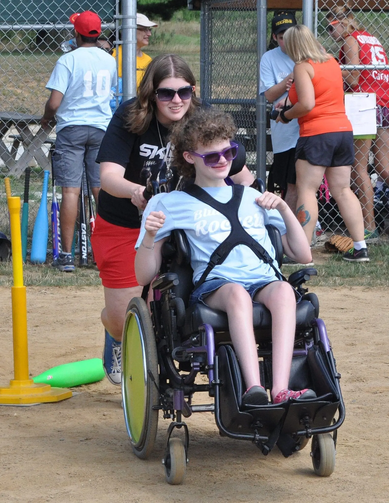 A young person in a wheelchair is being pushed by an older person, both wearing sunglasses.