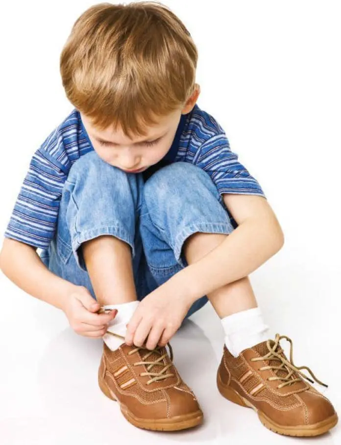 A young boy in jeans and a blue striped shirt ties his brown shoes.