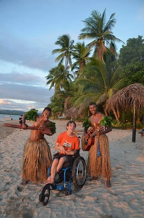 Julie Jones sits in a wheelchair on a beach between two men in grass skirts playing instruments.