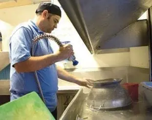 Man in blue shirt and black hat washing a large metal bowl with a spray nozzle.