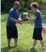 Two people in blue shirts and black shorts pass a medicine ball outdoors on a grassy field.