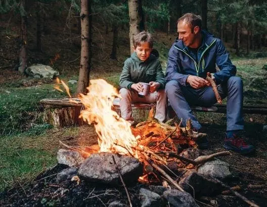 Man and boy sit by a campfire in the woods.
