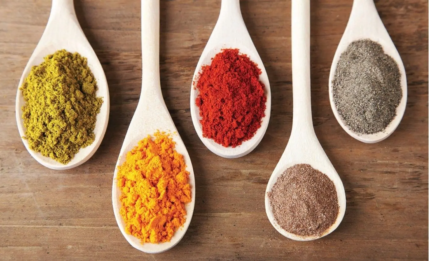Five ceramic spoons hold colorful spices on a wooden surface.