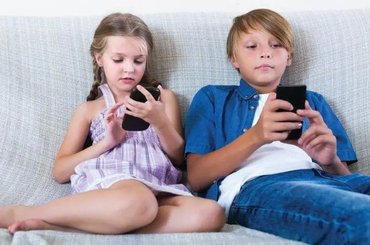 Two children sit on a couch looking at their smartphones.