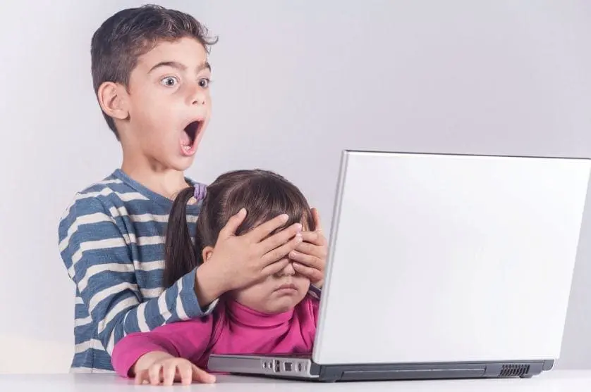 Two children react with shock and fear to a laptop screen, with the older child covering the younger child's eyes.