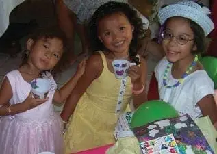 Three young girls in party dresses and hats hold teacups.
