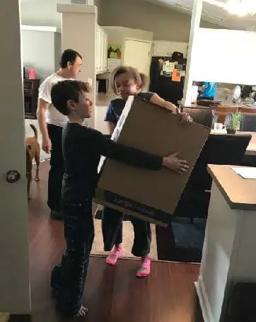 Two children carry a large box while a man stands in the background.