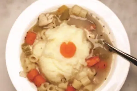Chicken noodle soup topped with a scoop of mashed potatoes and a carrot slice.