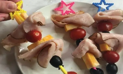 Hand holding a skewer made with folded deli meat, cubed cheese, and tomatoes topped with a star-shaped pick.
