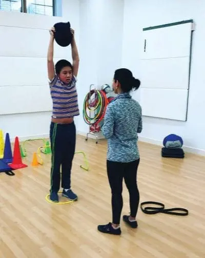 Boy holding kettlebell overhead while woman watches.
