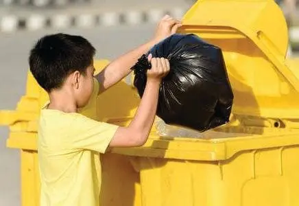 A young boy in a yellow shirt throws a full black trash bag into a large yellow dumpster.