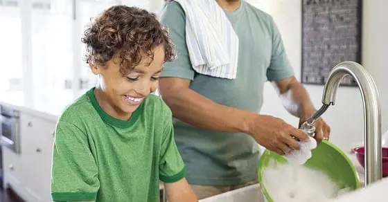Child smiling while washing dishes with an adult in the background.