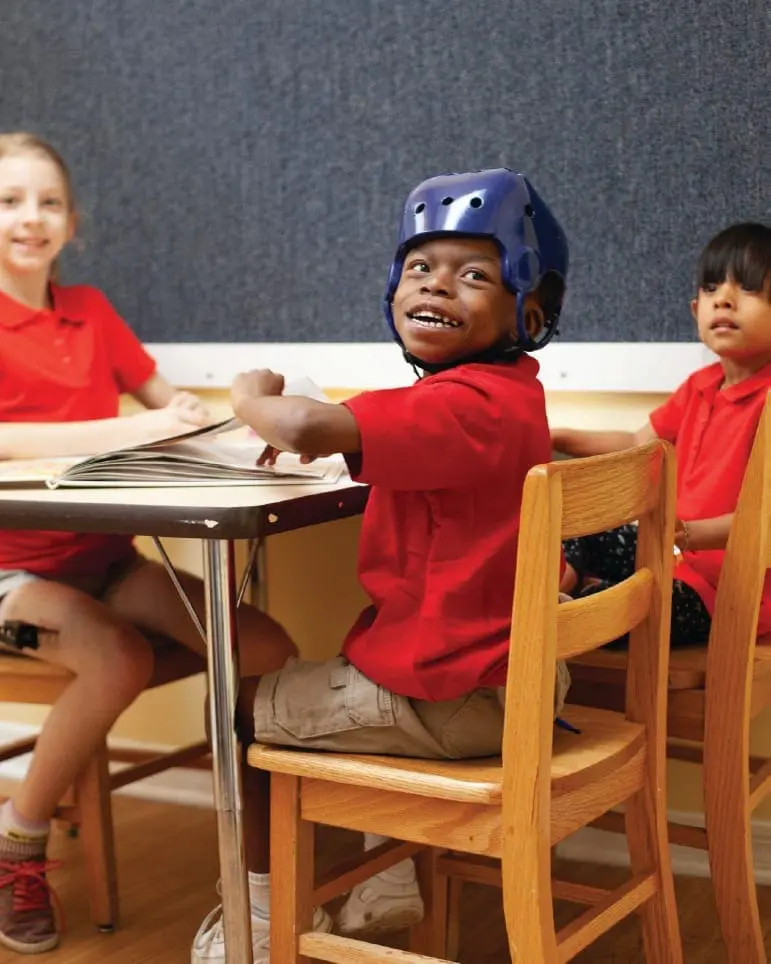 Three children in red shirts sit at a table, one wearing a blue helmet.