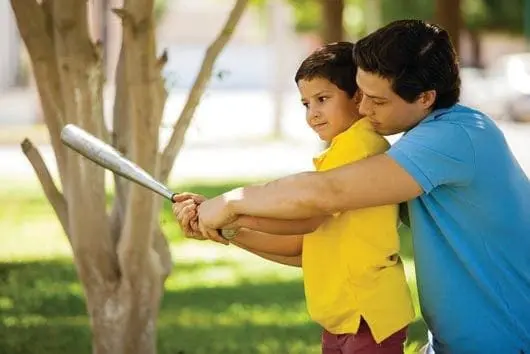 A man helps a boy swing a baseball bat outdoors.