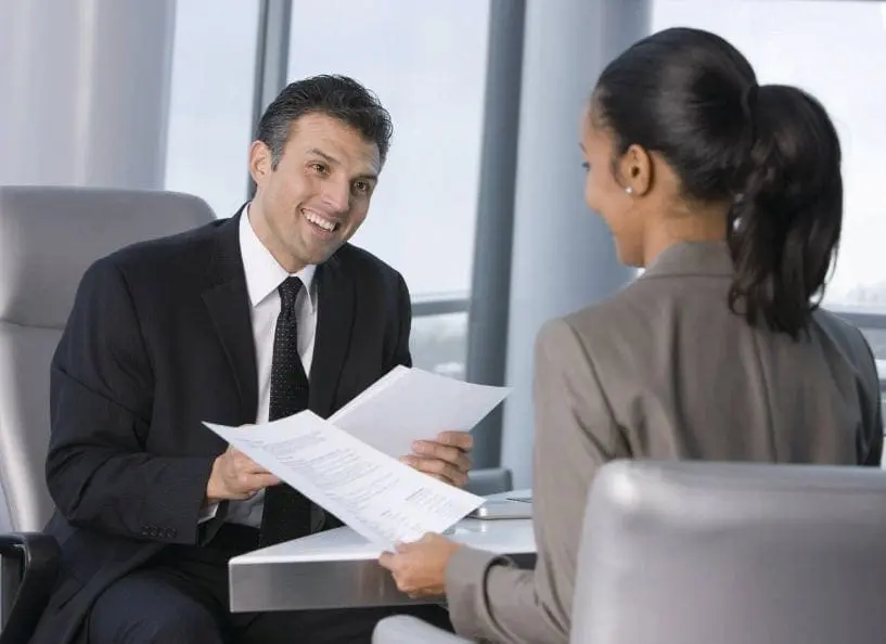 A man in a suit smiles while showing documents to a woman in a suit.