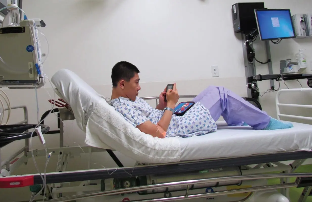 Jian plays a video game on a tablet while lying in a hospital bed.