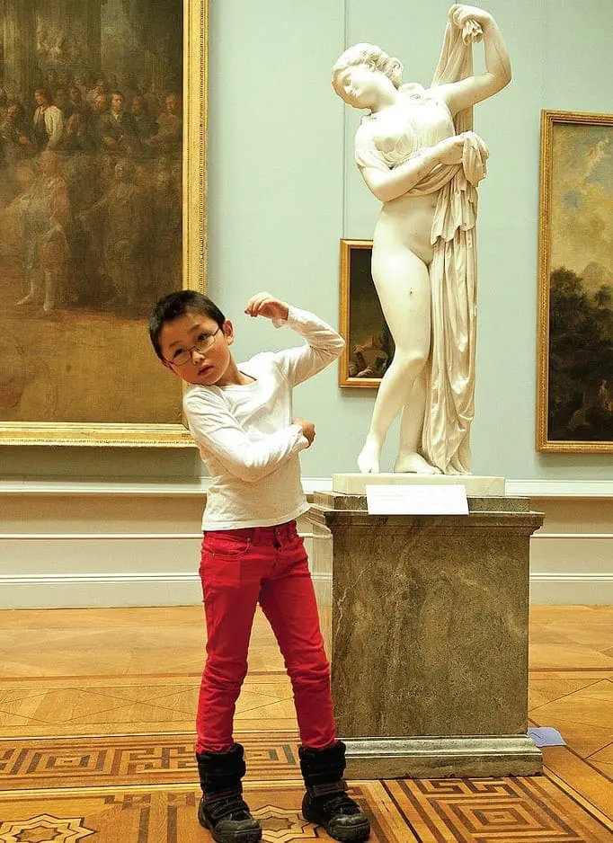 A young child strikes a pose next to a classical sculpture in a museum.