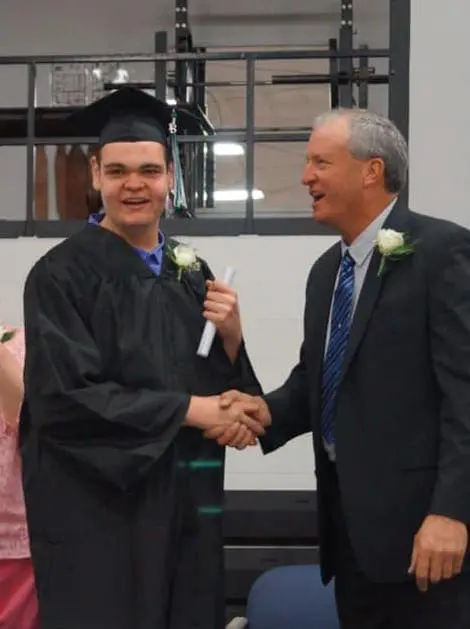 Two men shake hands, one in a graduation cap and gown, the other in a suit.