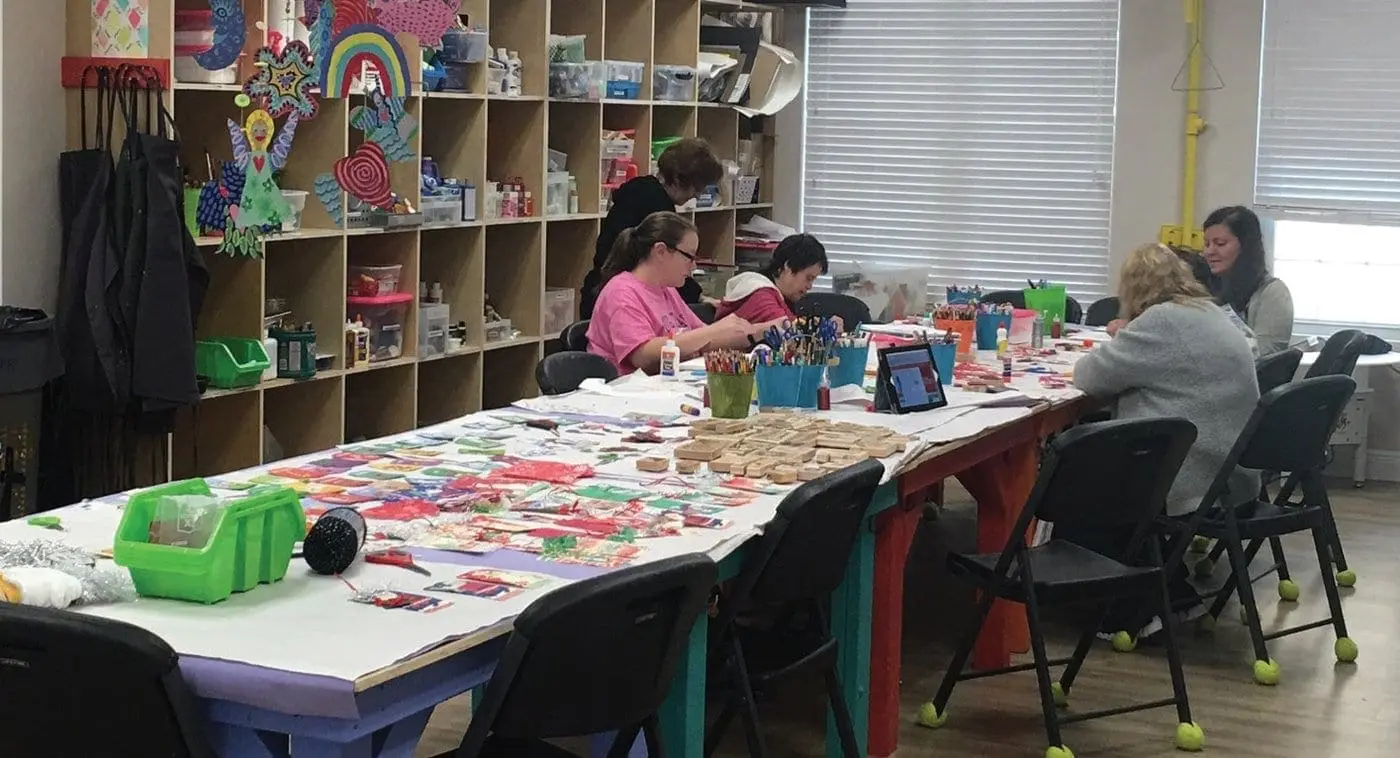 Several people sit at a long table in a room with shelves of supplies.