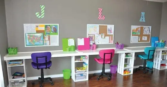 Three desks are set up along a wall with colorful chairs and organizational bins.